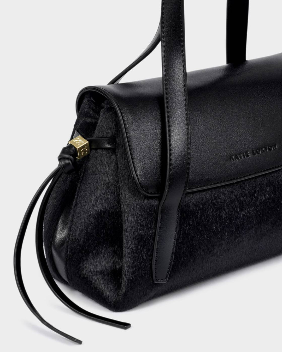 Alyssa Faux Pony Top Handle Bag in black vegan leather with fur accents, flap closure, gold drawstring detailing, “Katie Loxton” embossed on the side, and a crossbody strap. Sizes available: not specified.