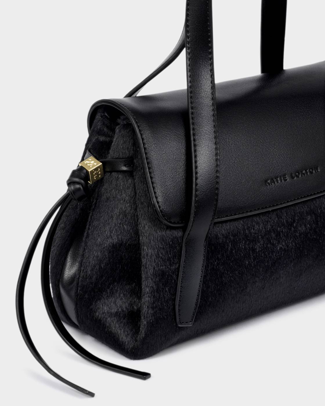 Alyssa Faux Pony Top Handle Bag in black vegan leather with fur accents, flap closure, gold drawstring detailing, “Katie Loxton” embossed on the side, and a crossbody strap. Sizes available: not specified.