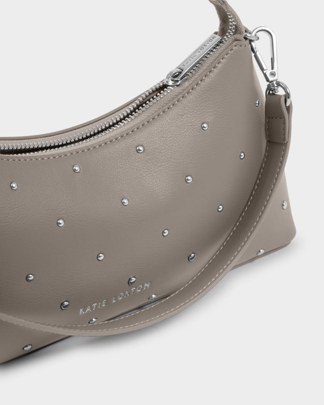 Taupe curved studded vegan leather handbag with silver studs, silver zipper, adjustable detachable strap, and “Katie Loxton” embossed in silver. Available in small size.