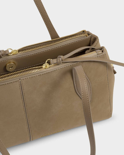 The Hattie Suedette Shoulder Bag in taupe vegan leather features gold zippers, double handles, exterior pockets, subtle branding, and a structured silhouette. Available sizes: Small, Medium, Large.