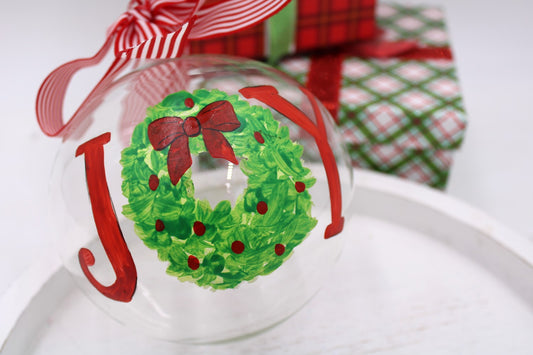 The 5" Hand-Painted Joy w/ Wreath Glass Ornament features "JOY" in red, with the "O" as a green wreath and red bow. Includes a red and white striped ribbon.