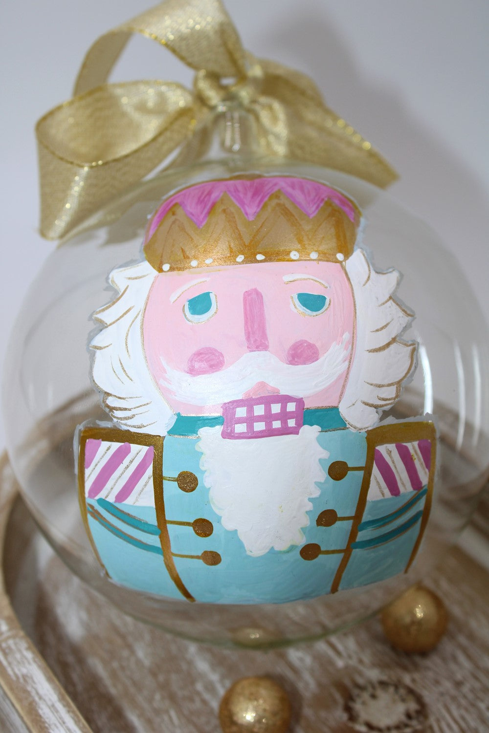 The 5" Hand-painted Nutcracker Brights Glass Ornament features a nutcracker with a pink face, white beard, blue uniform, and gold accents.