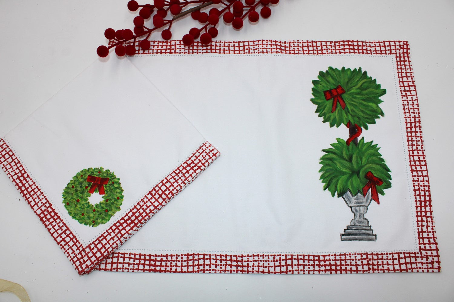 A set of four 18"x13" hand-painted topiary placemats with red bow motifs, accompanied by napkins featuring wreath designs and a red checkered border.