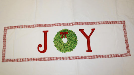 The 47"x14" Hand Painted Joy Table Runner features vibrant colors with "JOY" in red and green, a green wreath for the "O," against a white backdrop with a red and white checkered border. Available size: 47"x14".