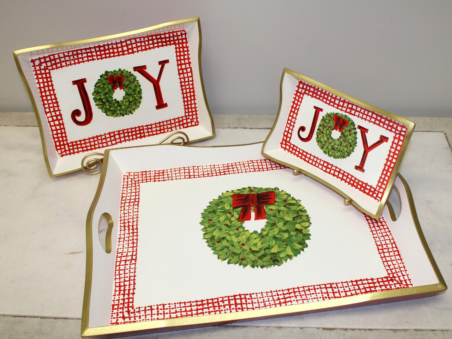 Handpainted Holiday Wood Trays feature a festive Christmas theme with "JOY" and wreath designs. Gold edges and red checkered borders enhance their seasonal charm. Available in various sizes.