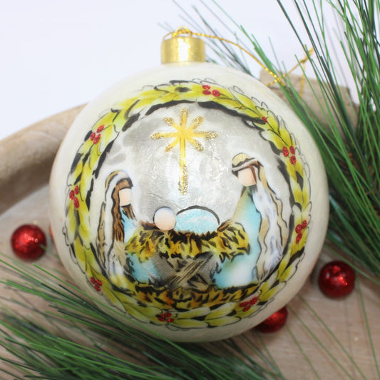 4" Handpainted Capiz Ball Ornament depicting the Holy Family in a nativity scene, enclosed by a holly wreath and topped with a golden star.