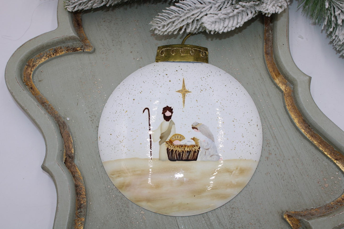 The 8"x.5"x10.5" Hand-Painted Neutral Nativity Metal Ornament showcases the Holy Family under a star, highlighted with gold speckles on a decorative green tray with pine branches.