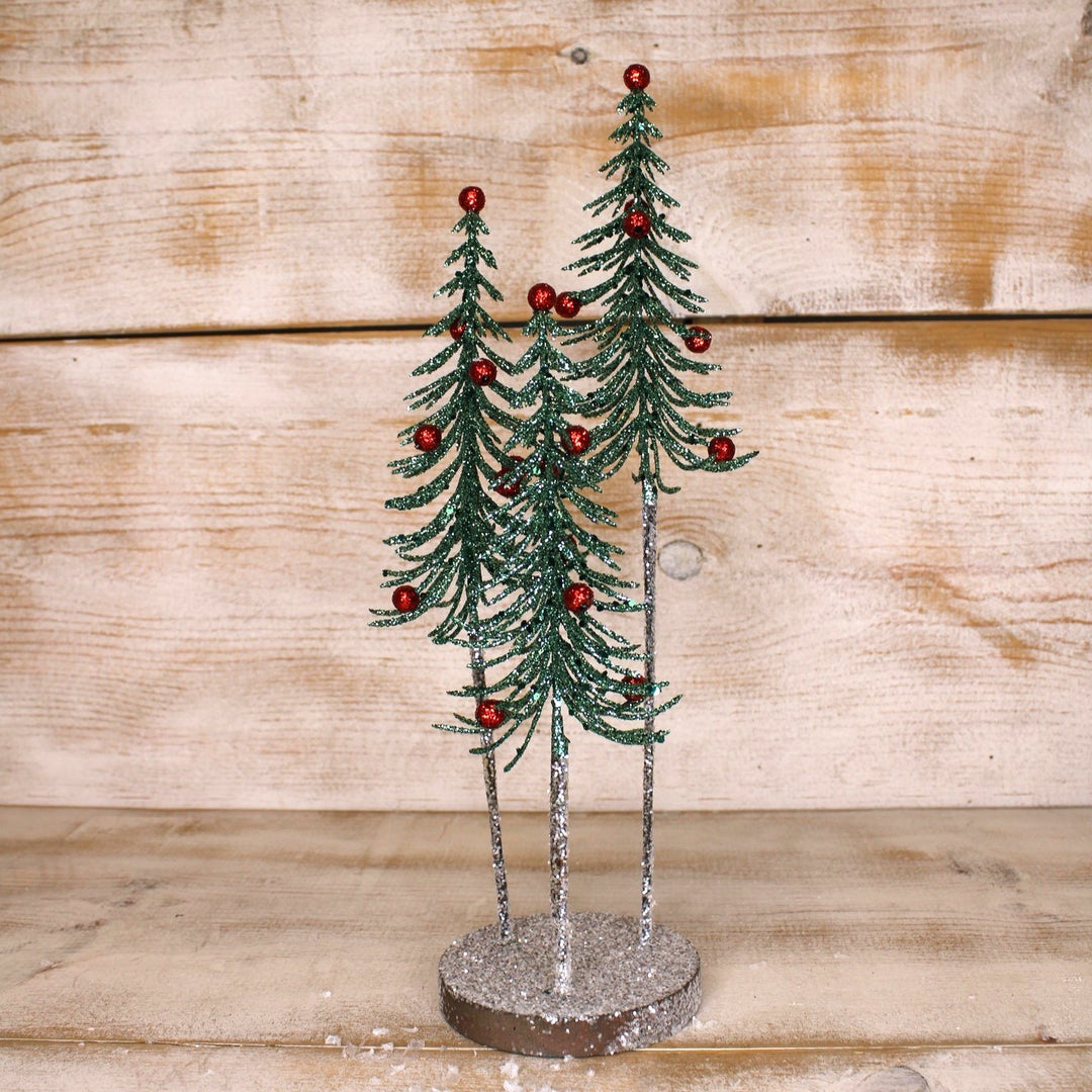 Three 5″x14″ glittery green trees with red berry accents on silver trunks and a round base.