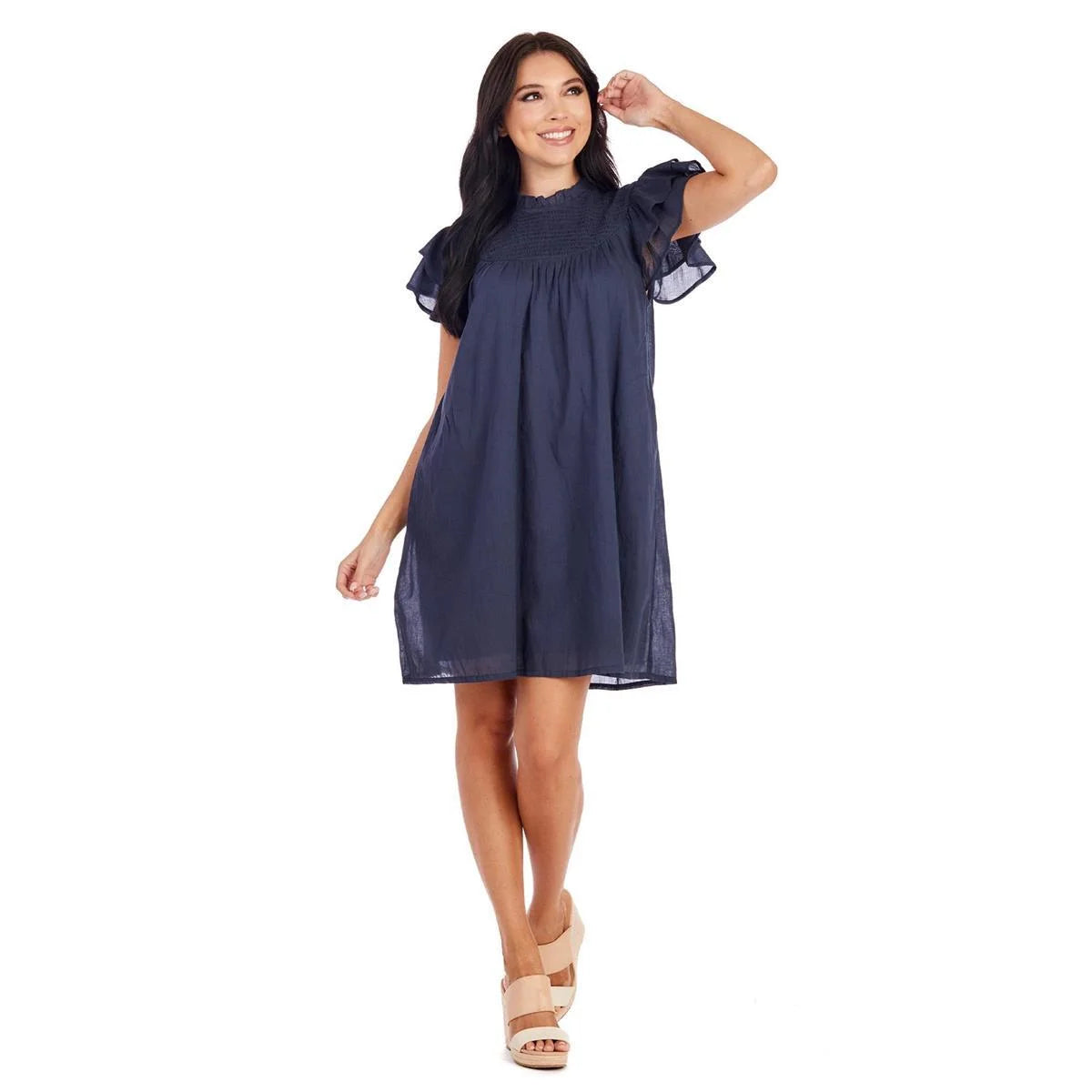 a room filled with vintage furniture. The Ashlyn Smocked Dress is available in small, medium and large sizes. This dress features ruffled sleeves, a knee-length hemline and a smocked chest design in dark blue color.