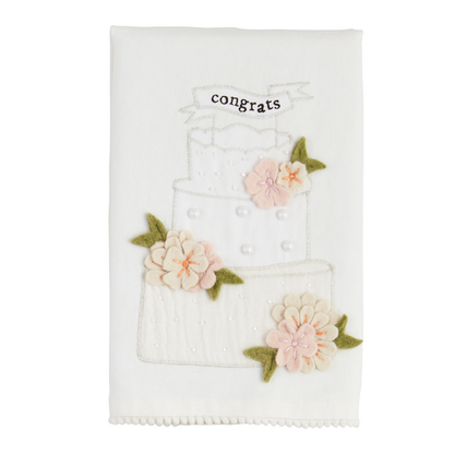 The Wedding Beaded Tea Towel is made of premium cotton and features a three-tiered cake with pink flowers, green leaves, and a "congrats" banner. It includes an embellished appliqué for added elegance. Size: Standard tea towel dimensions.