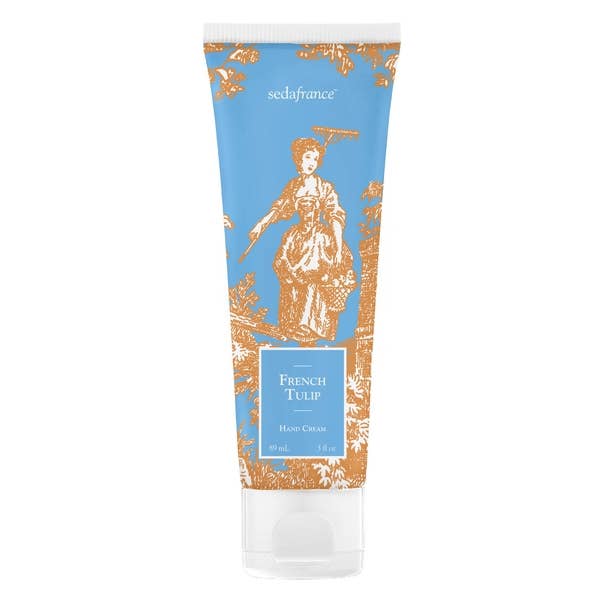 This is a tube of French Tulip Classic Toile hand cream. It displays a vintage-style illustration in orange and white hues depicting a woman dressed in traditional attire, amidst a garden setting.