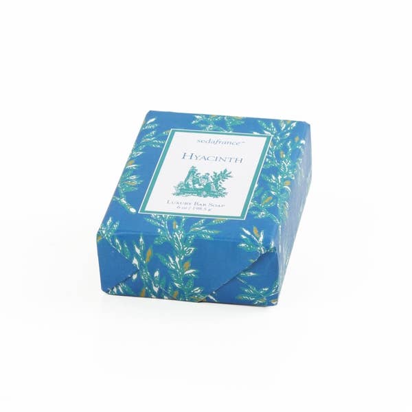 The product is a bar of Séduirence Hyacinth Luxury Face Soap. Wrapped in blue toile paper, the soap has a calming hyacinth fragrance. The dimensions are not specified. It is intended for facial use, offering luxury skincare with its hydrating formula and alluring scent. It comes in only one size and is presented on a pure white background for emphasis on purity and quality.