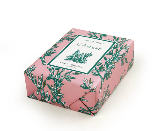 The A L'Ambre luxury musk toile soap comes with a distinctive and soothing musk scent. It is elegantly wrapped in a decorative pink and green floral-patterned paper that displays vintage-style labeling. The soap is designed for superior skin nourishment and gentle cleansing. It sits isolated on a white background, enhancing its visual appeal. This product is available in a standard size of 100g.