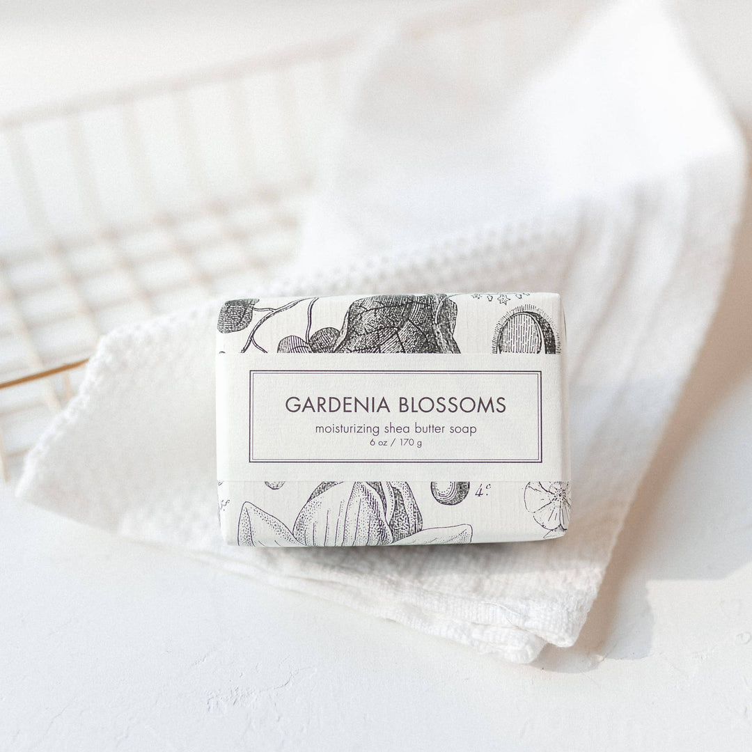 The Gardenia Blossoms Soap is a Bath Bar enveloped in a paper wrapper featuring a floral design. The soap offers a refreshing scent derived from gardenia blossoms. It boasts an aesthetic appeal with its rested placement on a textured white towel, providing it with clean and simple looks. The size of the product varies depending on availability.