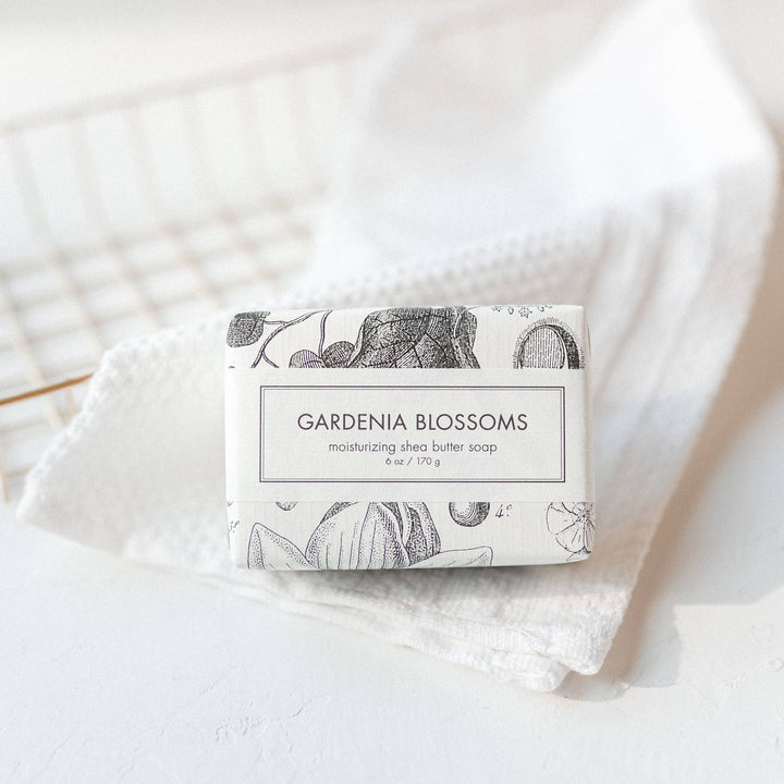 The Gardenia Blossoms Soap is a Bath Bar enveloped in a paper wrapper featuring a floral design. The soap offers a refreshing scent derived from gardenia blossoms. It boasts an aesthetic appeal with its rested placement on a textured white towel, providing it with clean and simple looks. The size of the product varies depending on availability.