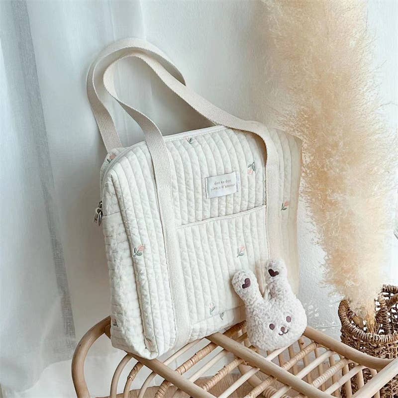 Embroidered diaper bag sitting on a woven end table with a plush bunny in front of it. Diaper bag is white with handles and embroidered pink and white flowers.