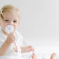 A toddler with blond hair sits against a white background, holding a BPA-free Pacifier Clip - Pink and Blue. The infant, wearing a white outfit, has a subtle smile and a clip in their hair.