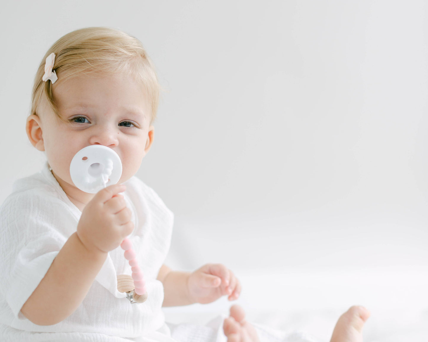 A toddler with blond hair sits against a white background, holding a BPA-free Pacifier Clip - Pink and Blue. The infant, wearing a white outfit, has a subtle smile and a clip in their hair.