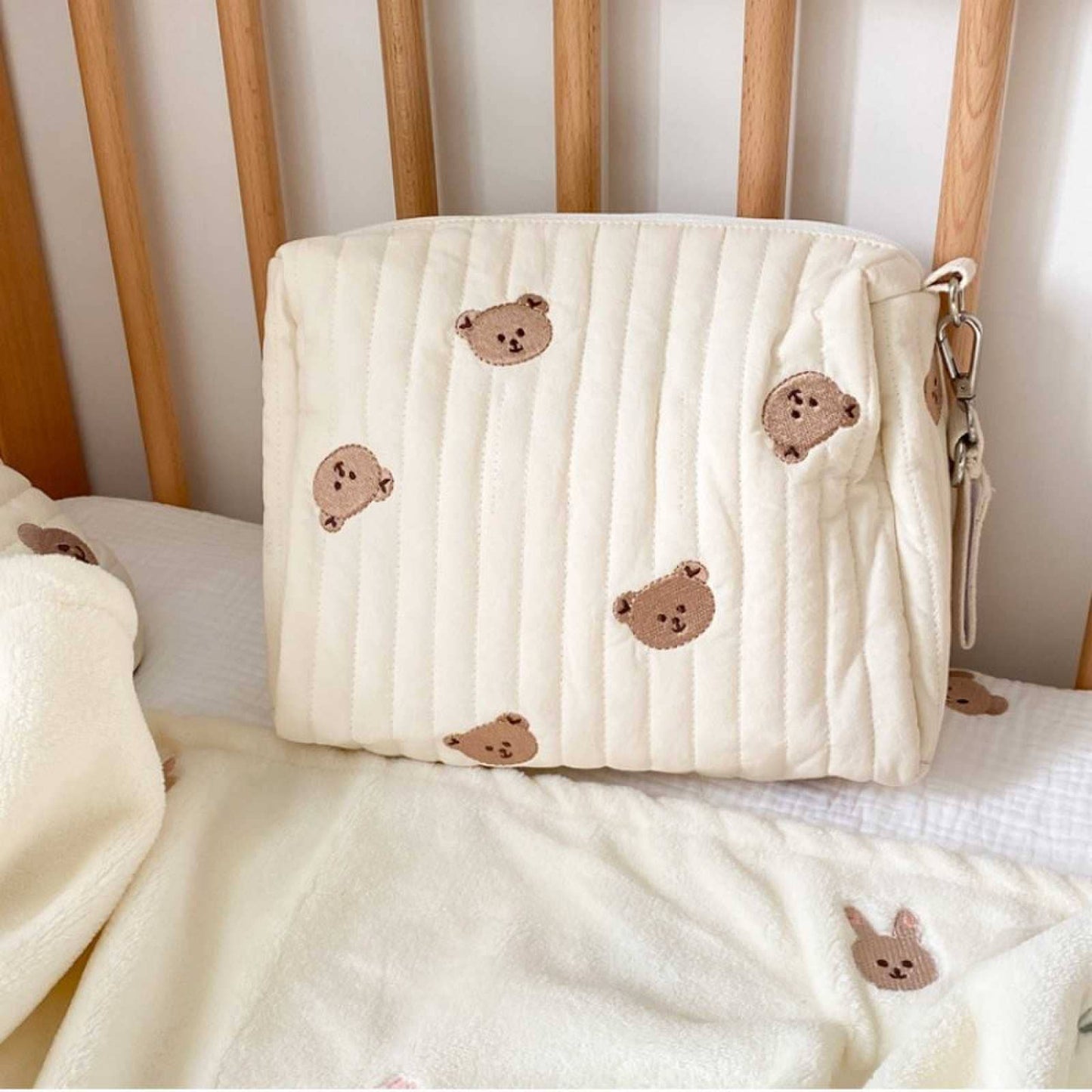 A plush cream-colored zippered pouch with an embroidered pattern of brown teddy bear faces, placed on a soft textured blanket in a cozy room setting, offering convenient storage options. Little Bear Embroidered Diaper Bag