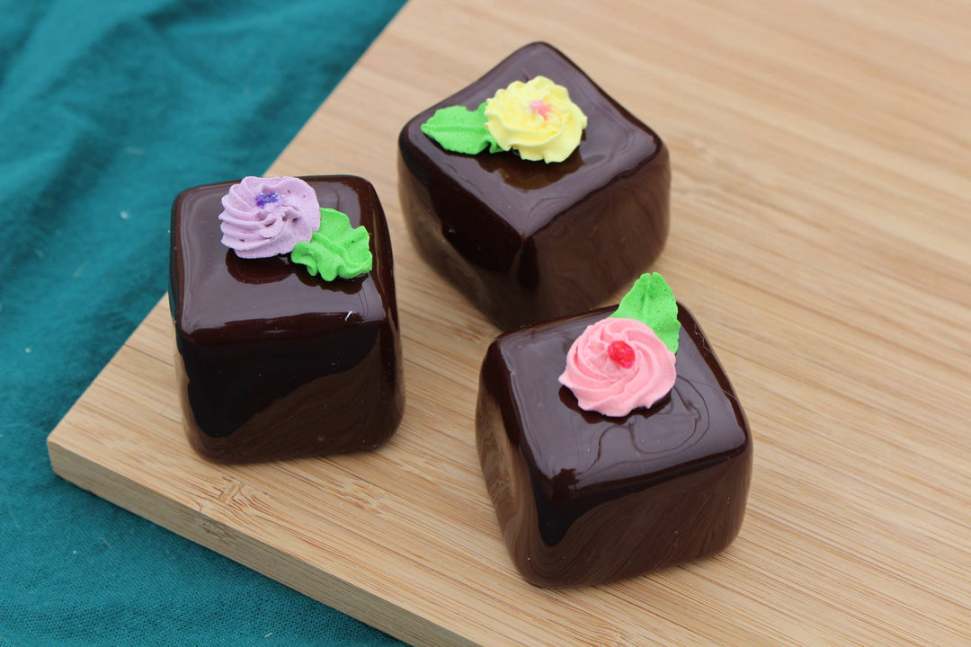 Three large glossy fake chocolate petit fours with icing flowers and green leaves; available in multiple sizes.