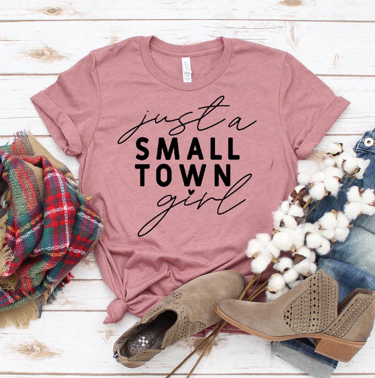 A pink Just A Small Town Girl Bella Canvas 3001 t-shirt with the phrase "just a small town girl" in black cursive text, paired with a colorful scarf, cotton branches, and brown ankle boots on a