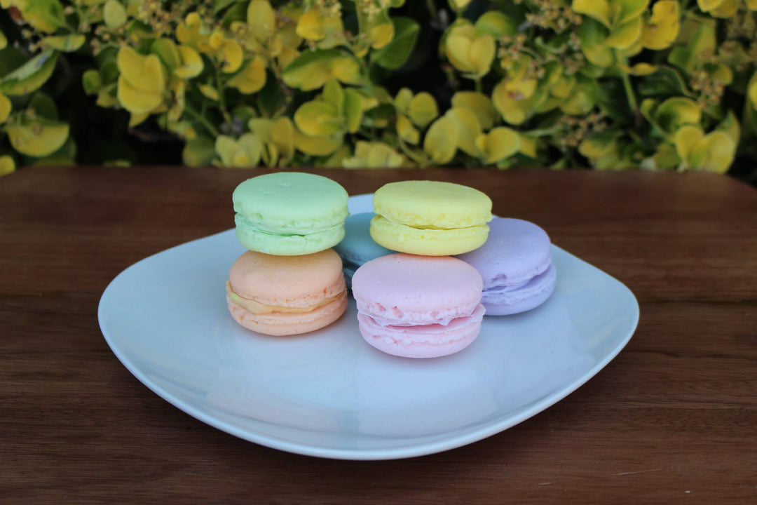 Five large fake macarons in green, yellow, orange, pink, and purple on a white plate; available in large size.