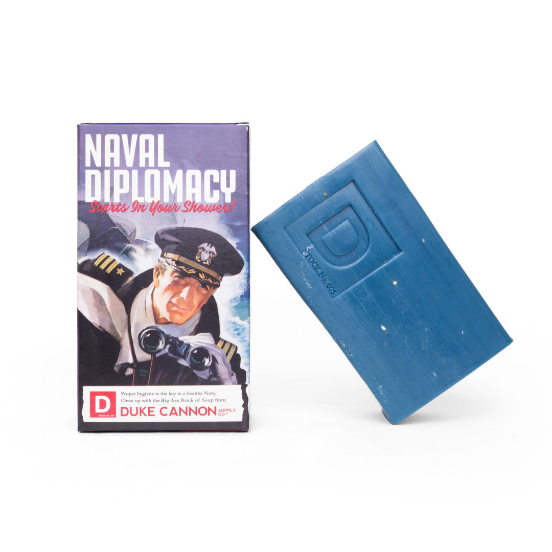 is a large, rectangular block in a deep blue color. The "Naval Diplomacy" fragrance is refreshing with notes of ocean breeze and leather. The soap measures 4.5" x 2.5" x 1.75", and weighs approximately nine ounces, making it three times the size of conventional soaps. It is made from natural ingredients with steel-cut grains for maximum grip and exfoliation features ensuring a thorough clean rinse without dryness or residue left behind on the skin after use. Comes sealed and packed in an artisan crafted