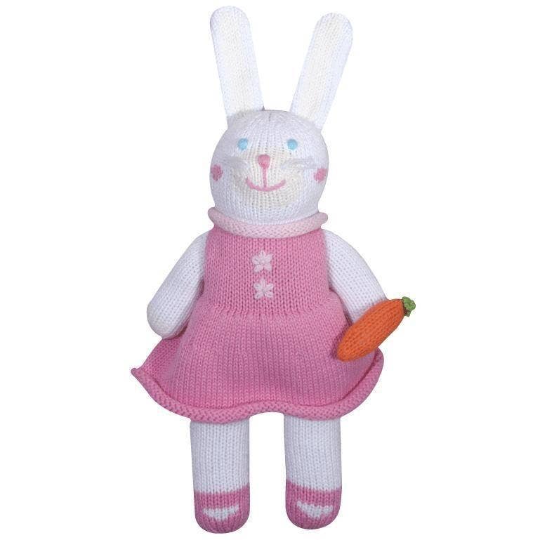 A Harriett the Bunny Knit Doll wearing a pink dress, standing upright and holding a small orange carrot. The bunny has white and pink striped legs and a cheerful expression.