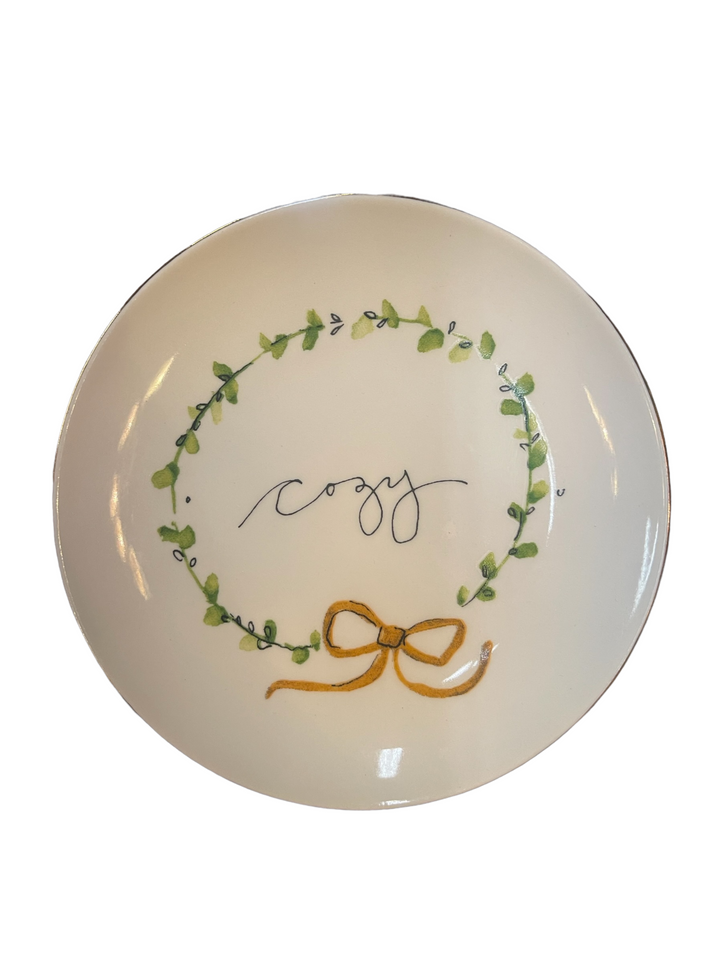 5” round white stoneware plate with green leaf wreath, “cozy” in cursive, and yellow bow at base.