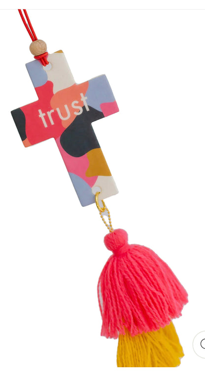This is a decorative cross featuring a colorful abstract design and the word "trust" imprinted on it. It dangles from a red string, with a sizeable pink tassel at the bottom, giving it an artistic flair. The product doubles as an air fresher, providing aesthetics and functionality in one item.