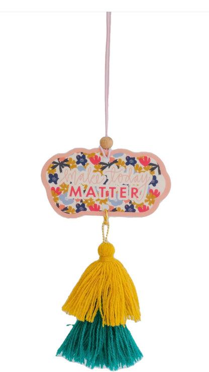 Fragranced car charm with a hanging sign featuring the phrase "make today matter!". Characterized by colorful flower embellishments, suspended on a pink string, and adorned with a yellow and green tassel. Available in one size.