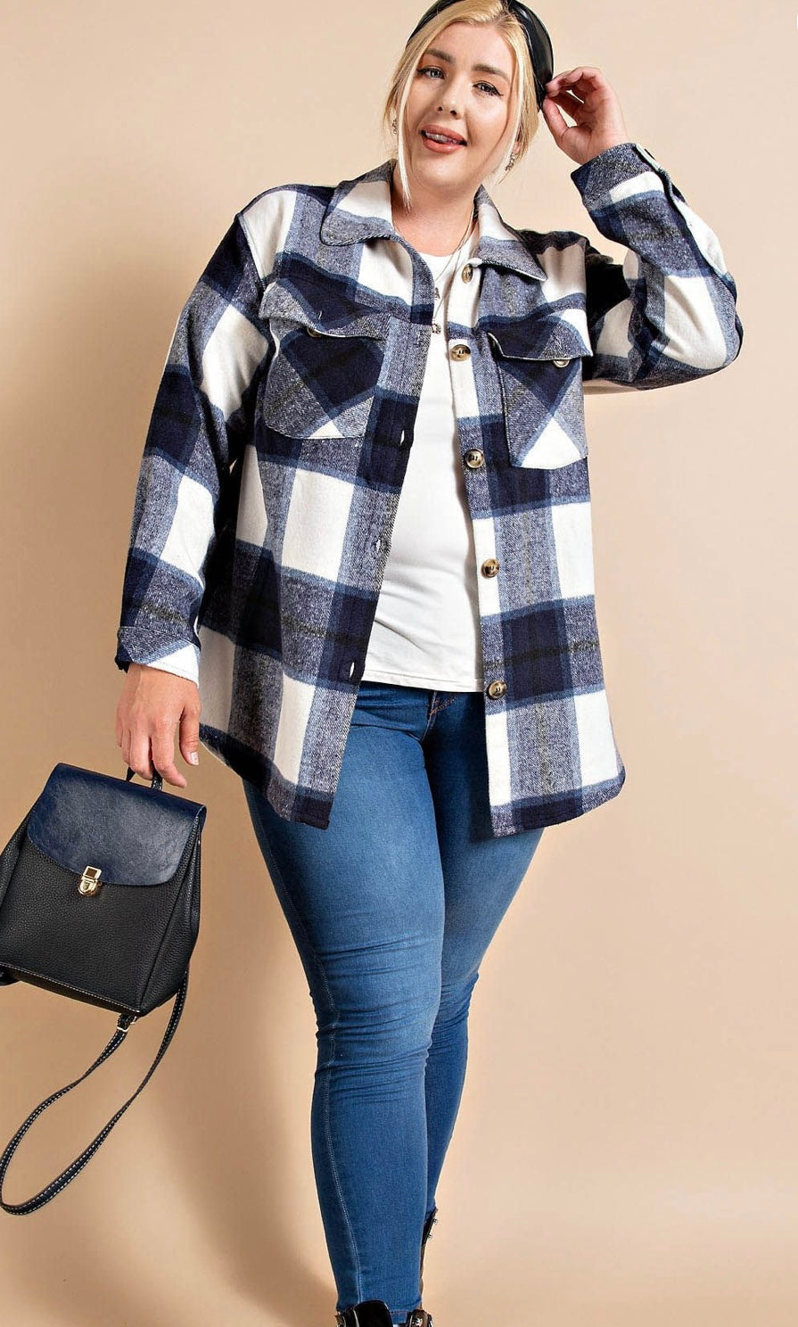 A woman in a Brushed Plaid Button Down Shirt - PLUS over a white top and blue jeans poses with a black handbag, touching the brim of her hat against a neutral background. The shirt is made from navy.