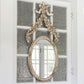 The Petite Flower Oval Mirror with antique white beaded frame and floral ribbon accent. Available in small and medium sizes.
