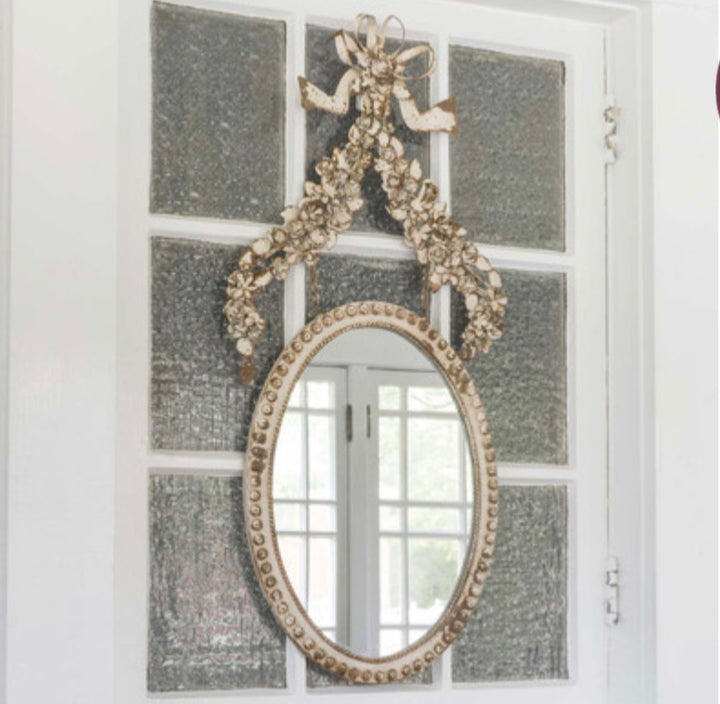 The Petite Flower Oval Mirror with antique white beaded frame and floral ribbon accent. Available in small and medium sizes.