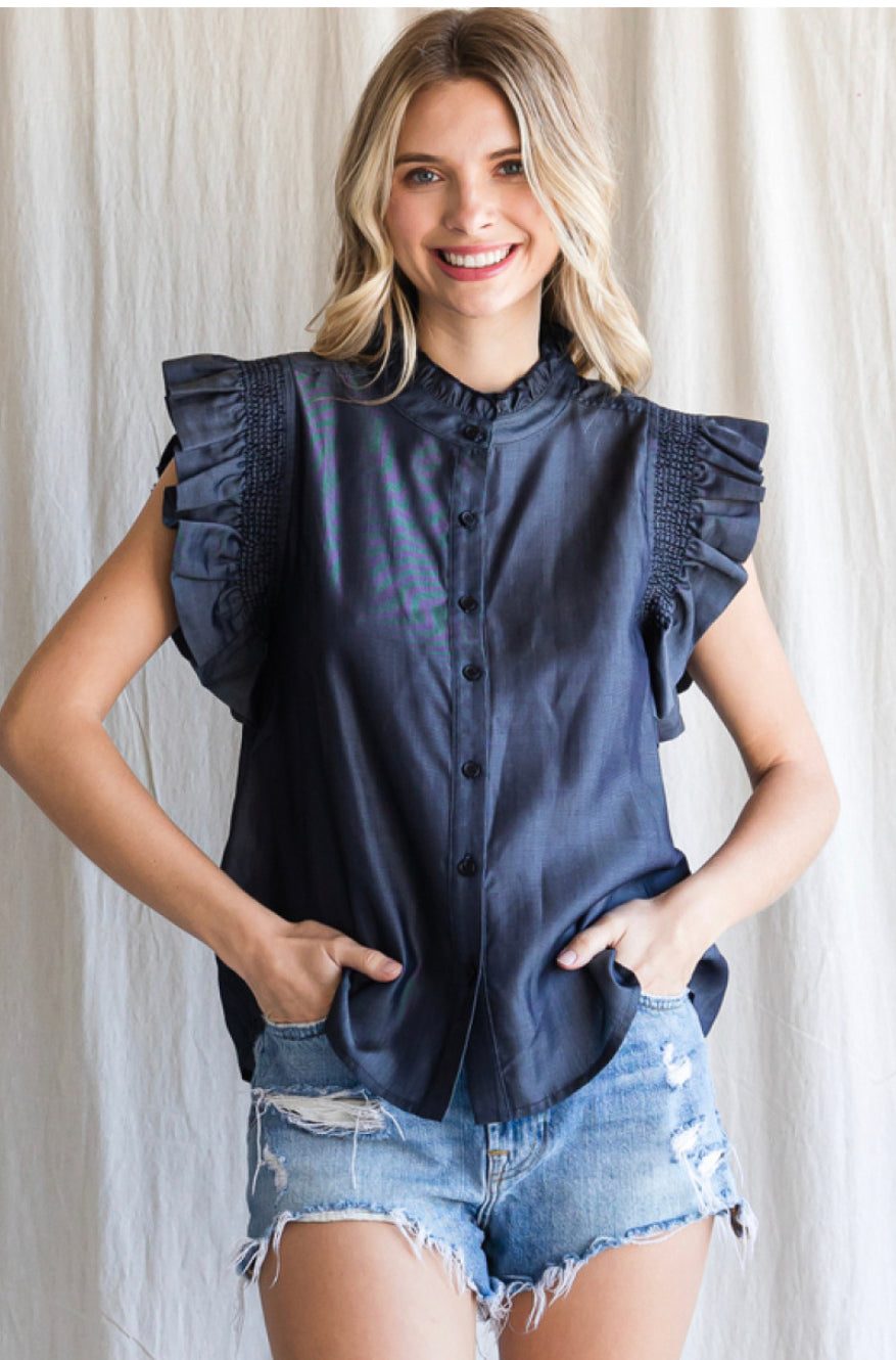 A smiling woman with blonde hair, wearing a dark ruffled sleeve blouse with smocked frilled shoulders and distressed Denim Frilled Top, standing against a plain light background.