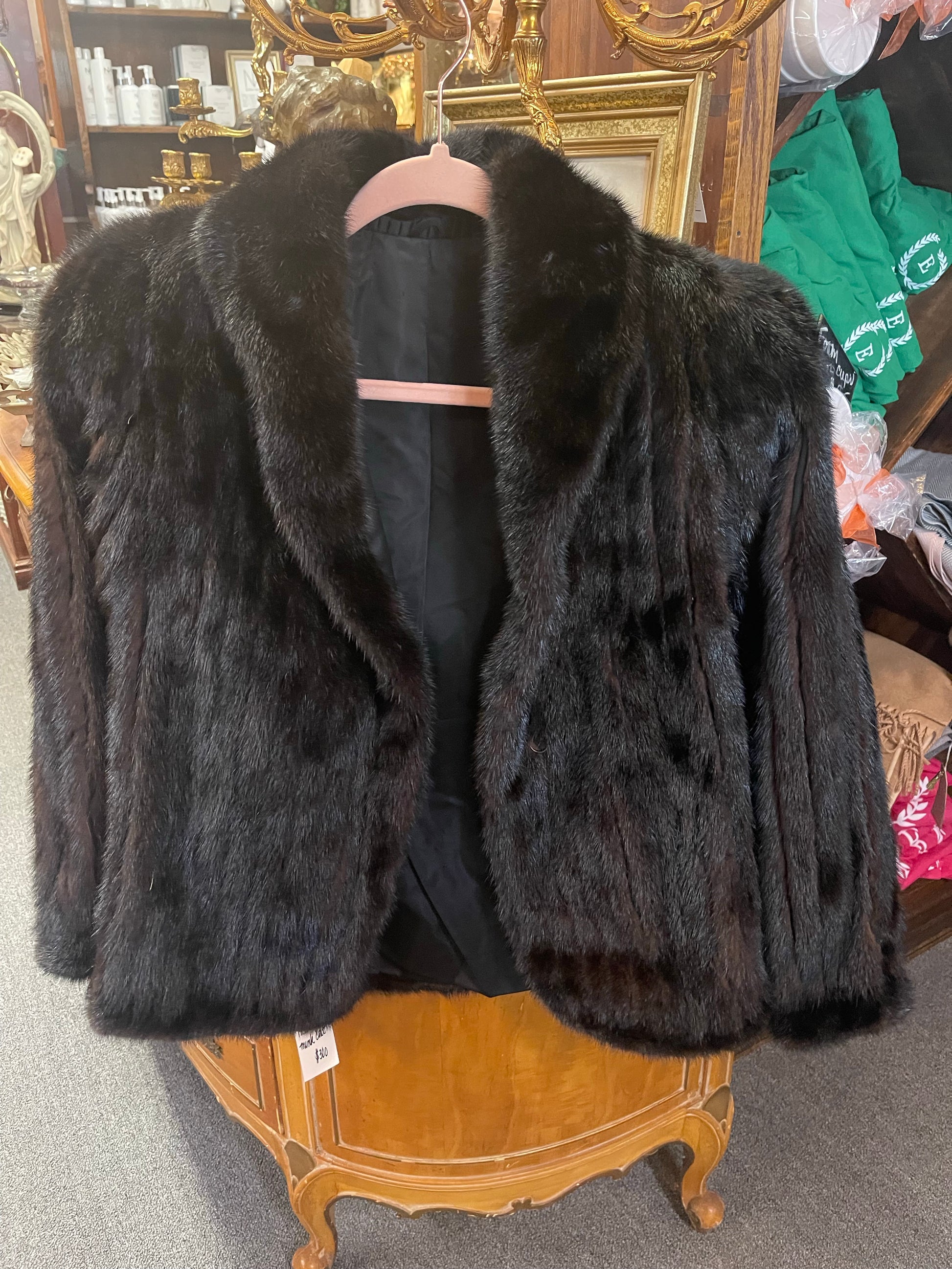 Black Short Mink Coat displayed on a wooden hanger, set against an ornately carved wooden dresser with various items and mirrors in the background.