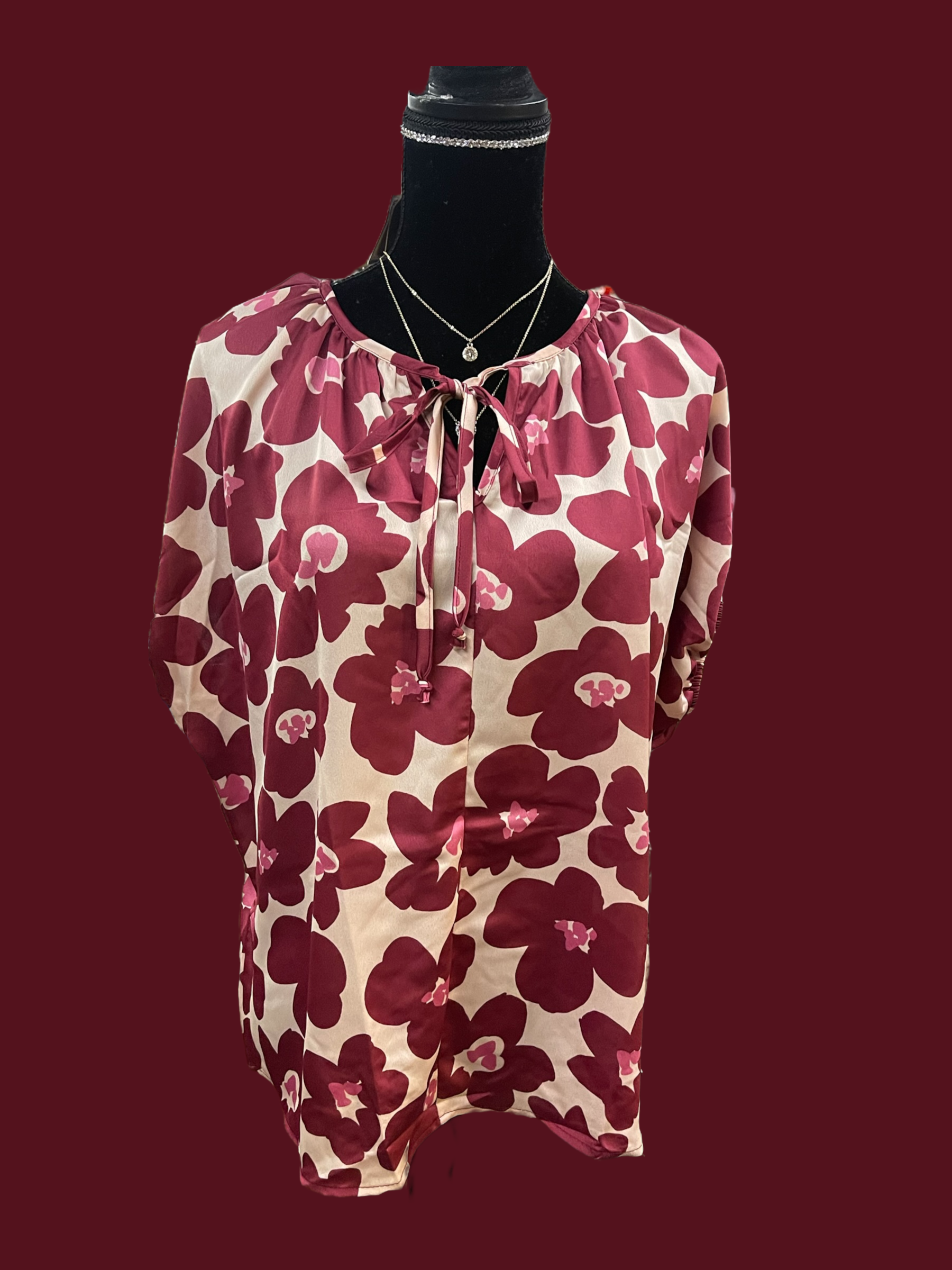 The Wine Floral Blouse available sizes include small, medium, large and extra-large. This blouse showcases a pink and red floral pattern with a bow-tied neckline. It is crafted from a silk-like fabric.