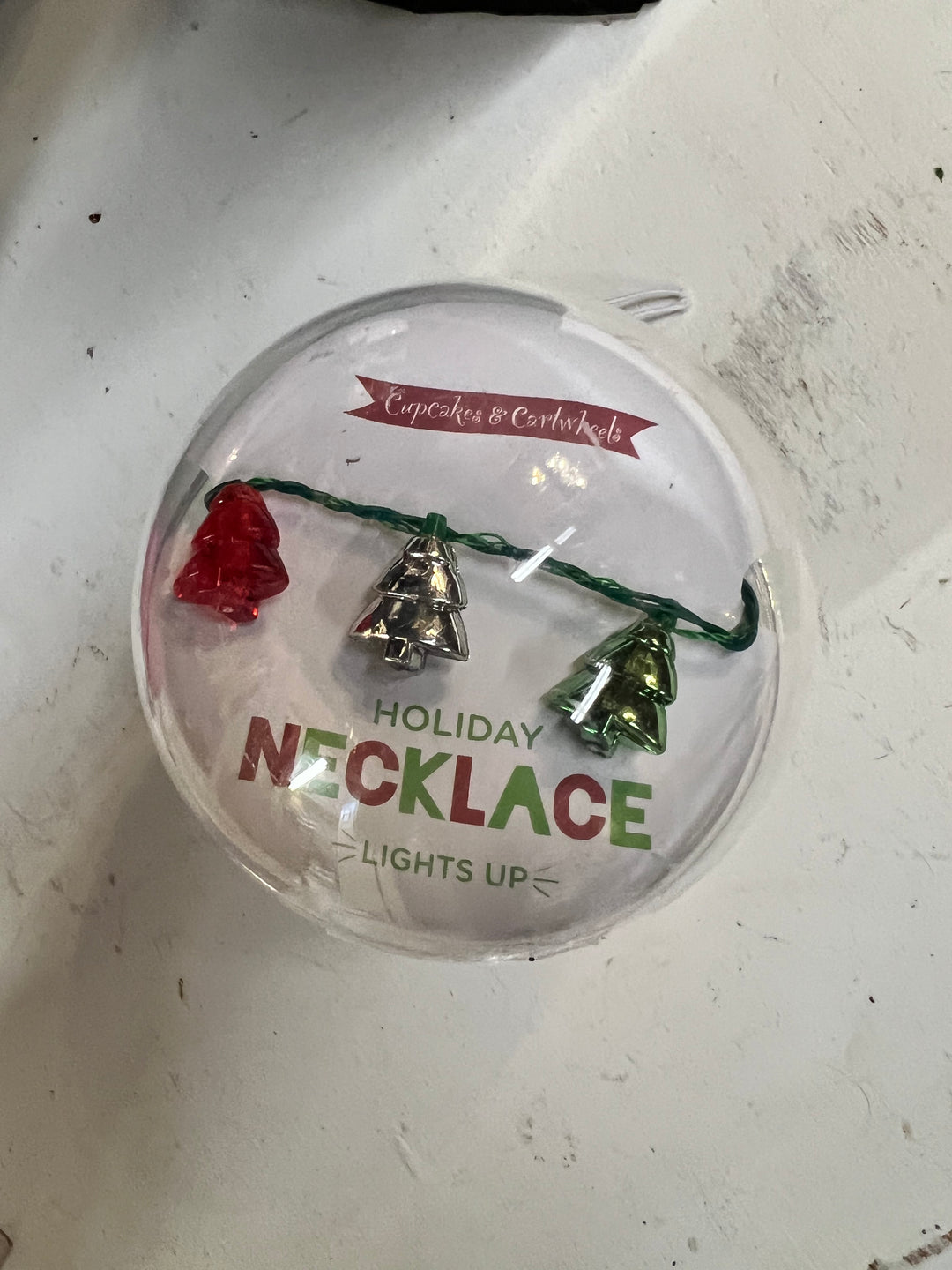 Holiday Light Up Necklace with red, silver, and green Christmas tree-shaped lights on a green wire, in a round plastic package.