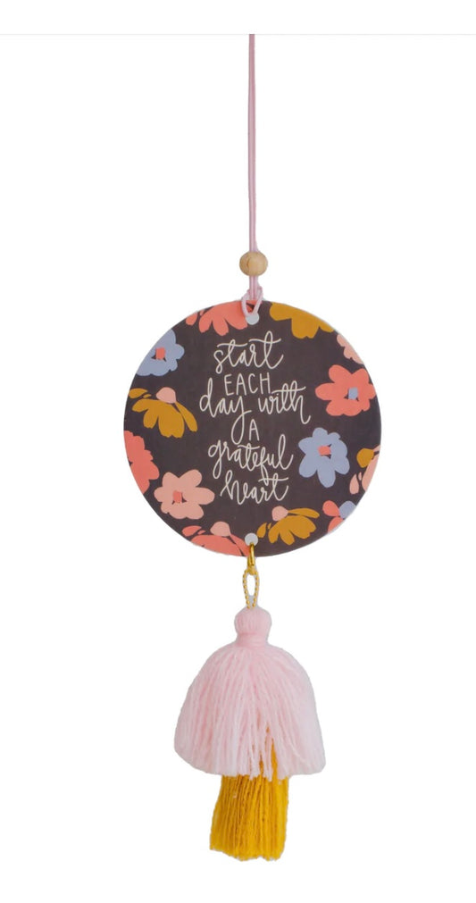 This product is a decorative hanging disc with a floral pattern. The text "start each day with a grateful heart" in cursive enhances the overall design. Embellished with pink tassel and yellow fringe, it doubles up as an Air Fresher. Available in one standard size.