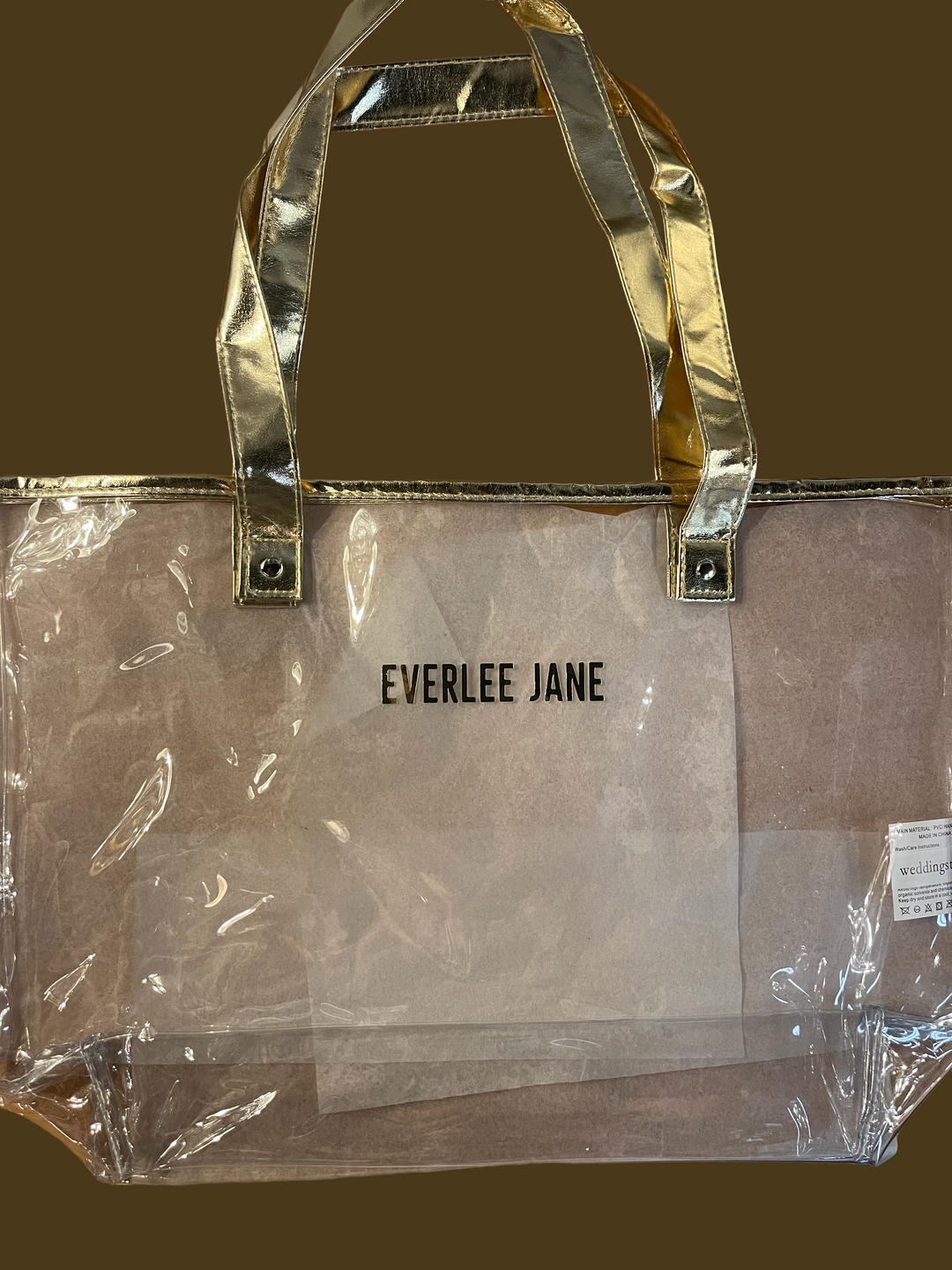 Clear tote bag with gold handles and trim, "EVERLEE JANE" on front. Stadium-approved. Sizes: Small, Medium, Large.