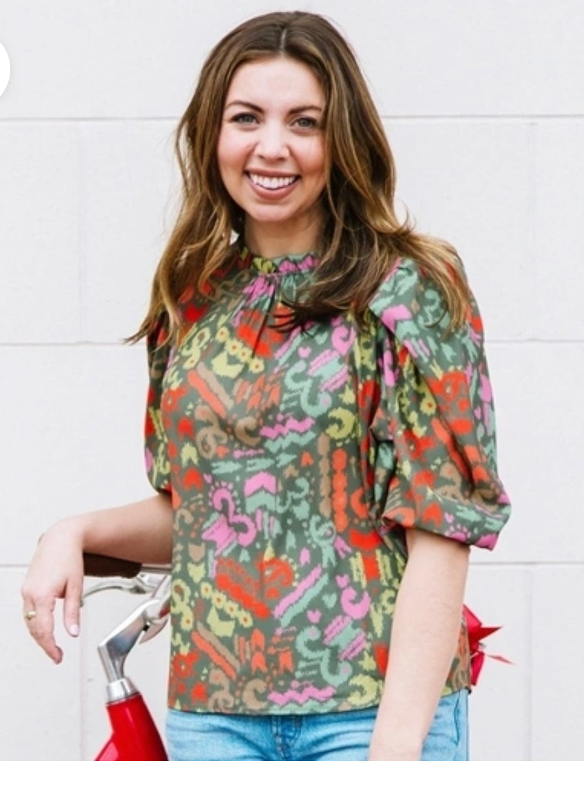 A smiling woman in a Pebble Path Olive Libby Top stands holding a bicycle pump, sizing it appropriately.