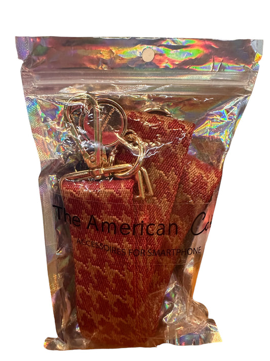 This product is a crossbody phone strap featuring a red and brown houndstooth pattern. Adjustable for versatile wear options, it includes both a metal keyring and crossbody straps. Packaged with care, the strap comes delivered in a distinct holographic zippered bag that bears "The American Case" label. The adjustable sizing of the straps makes it suitable for everyone. Please note that no other products are included other than the stated phone strap within its labeled packaging bag.