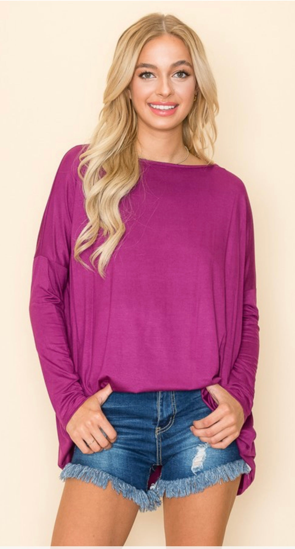 A young woman with blonde hair, smiling, wearing a A.gain “The Perfect Shirt” Long Sleeve Regular made of buttery soft Rayon Spandex in purple, and blue denim shorts, standing against a cream background.