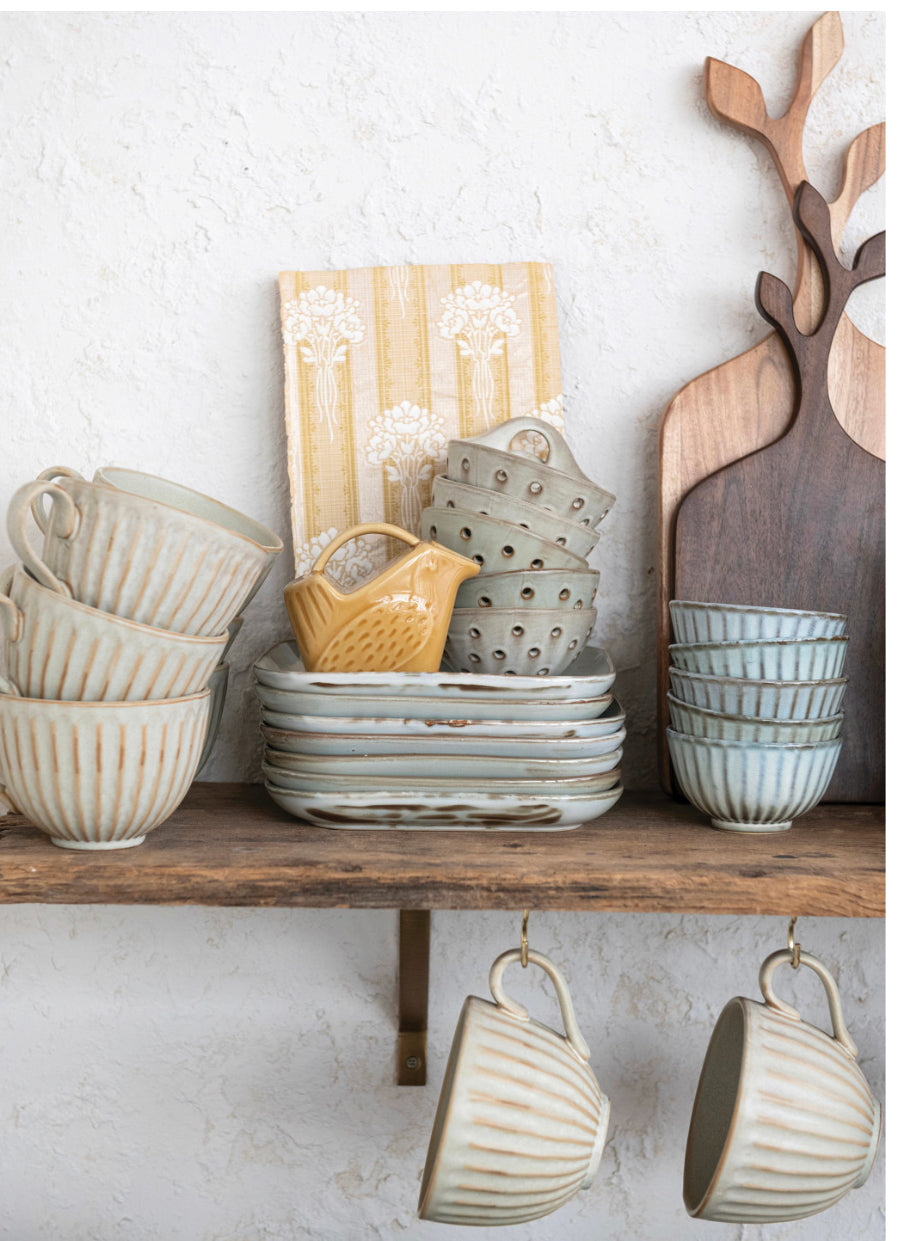 A rustic kitchen shelf in traditional design, fitted with a selection of ceramic tableware. The set includes striped mugs & plates, a colander and various sized bowls. Also featured is a unique 5 oz. stoneware bird shaped creamer available in three distinct colors. The set is complemented by a robust wooden cutting board. Mounted ideally against white walls, the product comes in one standard size.