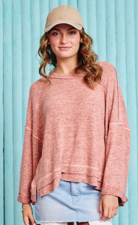 A young woman wearing a Dew Loose Fit Boat Neck Top in pink and light denim skirt, topped with a beige cap, standing against a blue striped backdrop. She has soft curls and is looking directly at the camera.