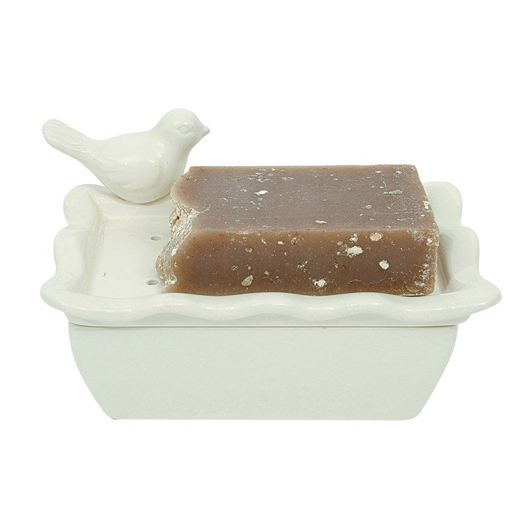 A beige bar of soap featuring fine salt flecks. Paired with a ceramic soap dish shaped like a bird bathtub, complete with a perched ceramic bird on the rim. Soap and dish are singular items for purchase only.